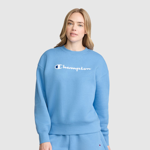 A woman wearing a light blue Champion Powerblend Crewneck Sweatshirt, Script Logo, and matching sweatpants stands facing forward against a plain light gray background.