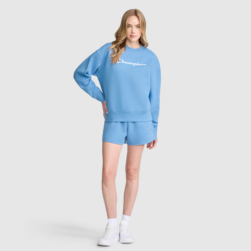 A person stands wearing a light blue Champion Powerblend Crewneck Sweatshirt with the script logo and matching shorts, paired with white sneakers. The background is plain light gray.