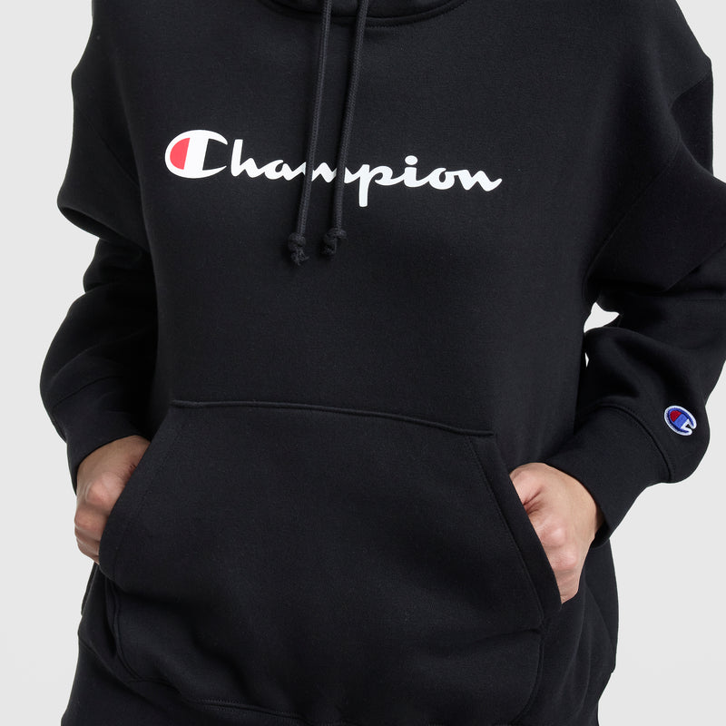 A person wears a black Champion Powerblend Relaxed Hoodie with hands in the front pocket. The chest and sleeve feature the white Champion logo with a red and blue "C." The background is plain and light-colored.