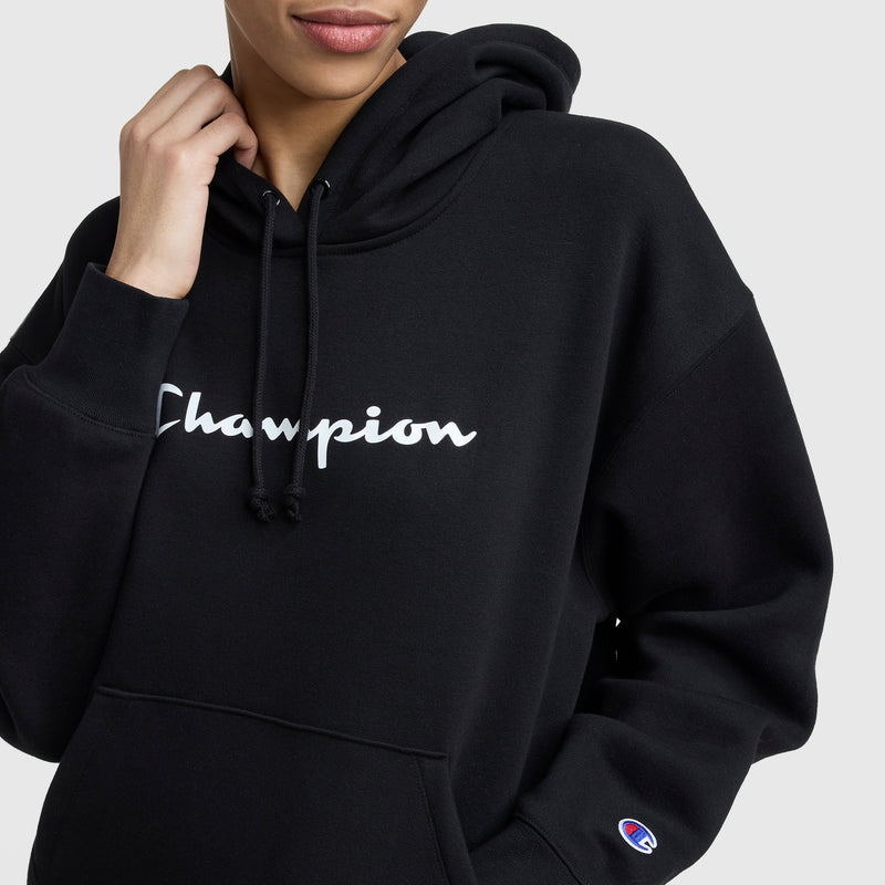 A person wearing a black Champion Powerblend Relaxed Hoodie with the hood up, displaying the Champion logo on the chest and sleeve, is touching the hood string and smiling slightly.