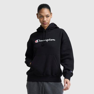A woman in a black Champion Powerblend Relaxed Hoodie with white and red logo text stands with one hand in her pocket, looking slightly to the side against a plain light gray background.