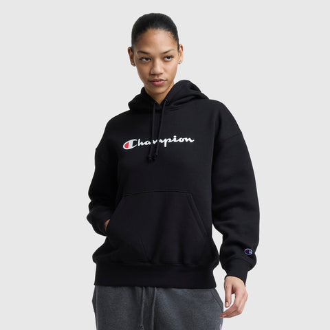 A woman in a black Champion Powerblend Relaxed Hoodie with white and red logo text stands with one hand in her pocket, looking slightly to the side against a plain light gray background.