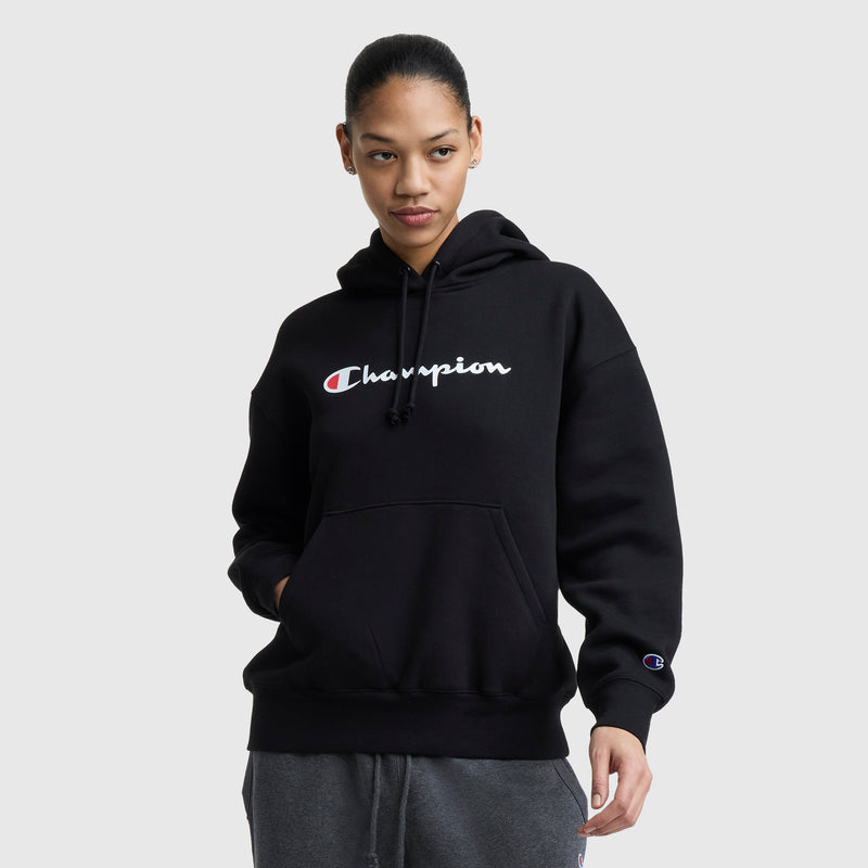 A woman in a black Champion Powerblend Relaxed Hoodie with white and red logo text stands with one hand in her pocket, looking slightly to the side against a plain light gray background.