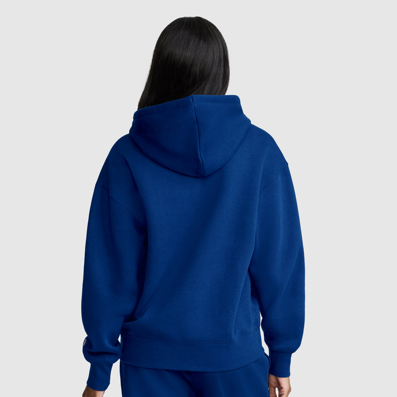 A person with long dark hair, facing away from the camera against a plain light gray background, wears a Champion Women’s Powerblend Relaxed Hoodie, Script Logo and matching blue pants. The midweight fleece of this Champion hoodie provides both comfort and style.