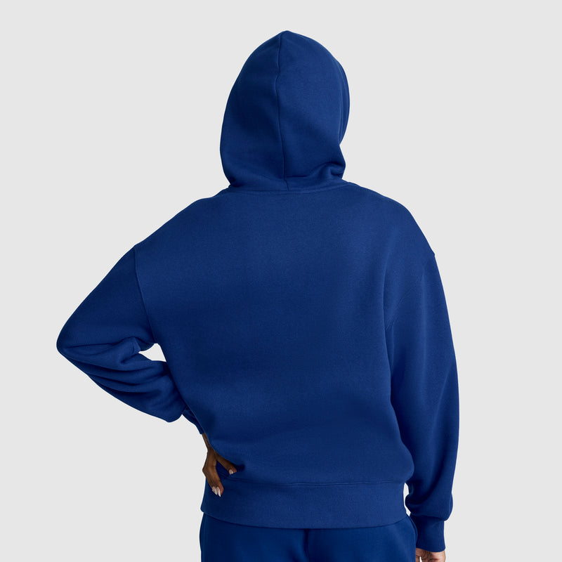 A person wearing a royal blue Champion Women’s Powerblend Relaxed Hoodie with a script logo and matching pants stands with their back to the camera, one hand on their hip, against a plain light gray background. The midweight fleece of the hoodie from Champion adds extra comfort and style to the outfit.