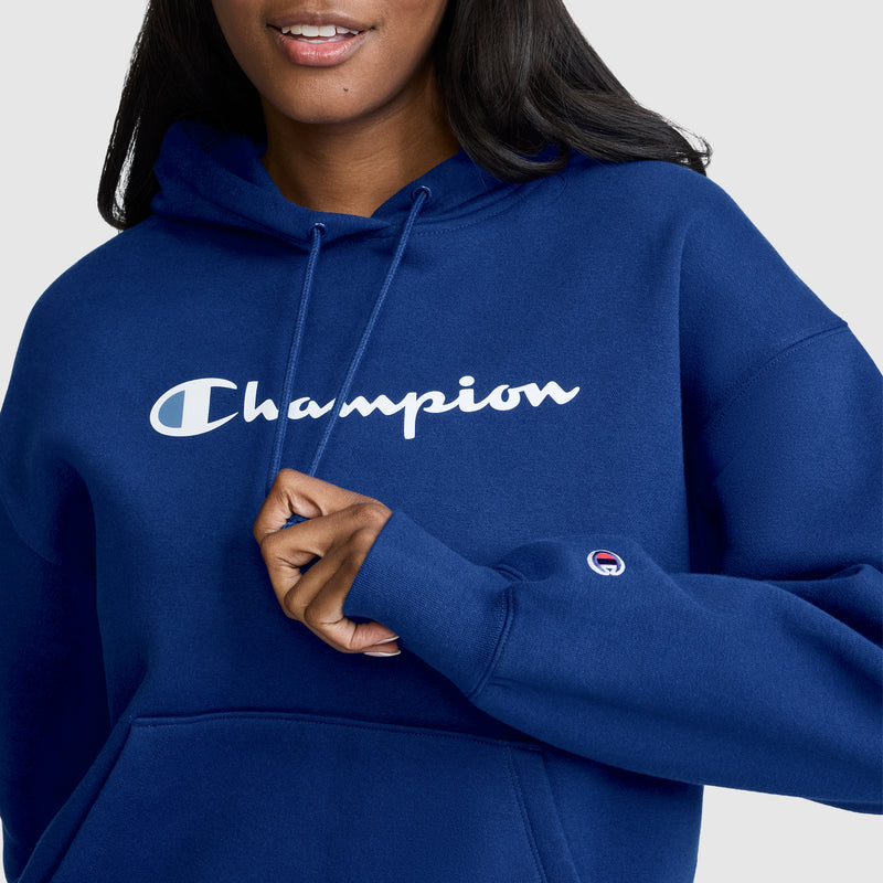 A person wearing a blue Champion Women’s Powerblend Relaxed Hoodie, Script Logo holds the hood’s drawstring. This women’s hoodie by Champion features the script logo printed in white across the chest and is crafted from midweight fleece for added comfort.