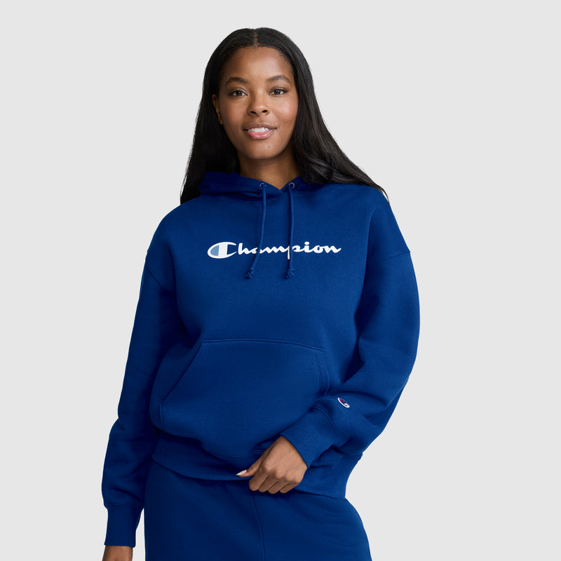 A woman wearing a blue Champion Women’s Powerblend Relaxed Hoodie with a script logo stands against a plain light gray background, smiling and looking at the camera. The midweight fleece of this Champion hoodie offers both comfort and style.