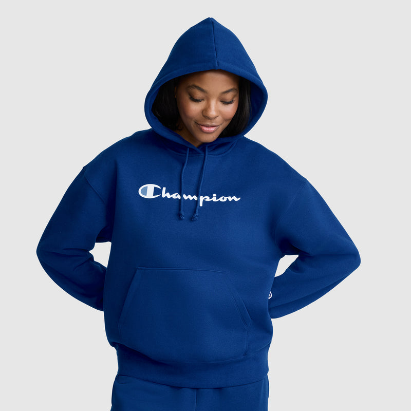 A woman in a blue Champion Women’s Powerblend Relaxed Hoodie, featuring the iconic script logo on the front, stands with her hands behind her back, looking down and smiling. This Champion hoodie is crafted from soft midweight fleece for ultimate comfort.