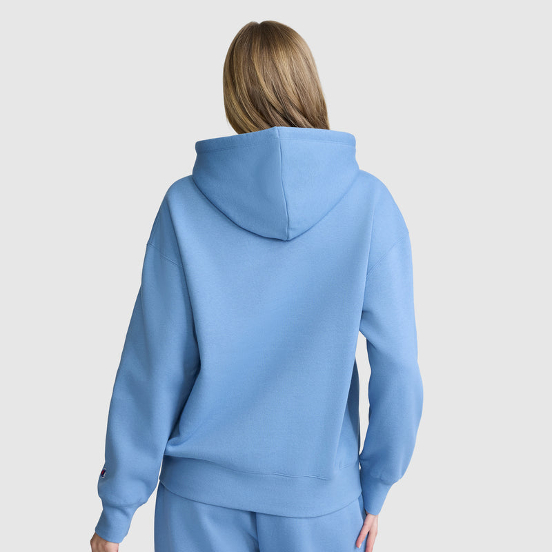 A person with long light brown hair is shown from the back, wearing a Champion Women’s Powerblend Relaxed Hoodie with a script logo and matching fleece sweatpants against a plain light background.