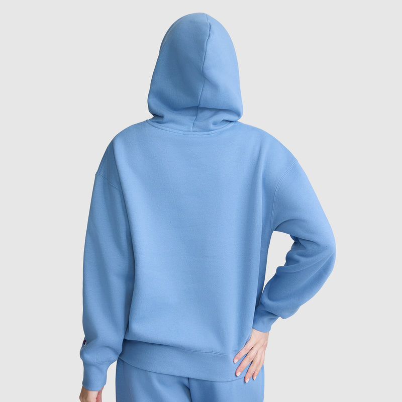 A person is seen from behind wearing the Champion Women’s Powerblend Relaxed Hoodie, Script Logo by Champion, paired with matching light blue pants. The hood is up, concealing their head and face, and they have one hand on their hip. The background is a plain light gray.