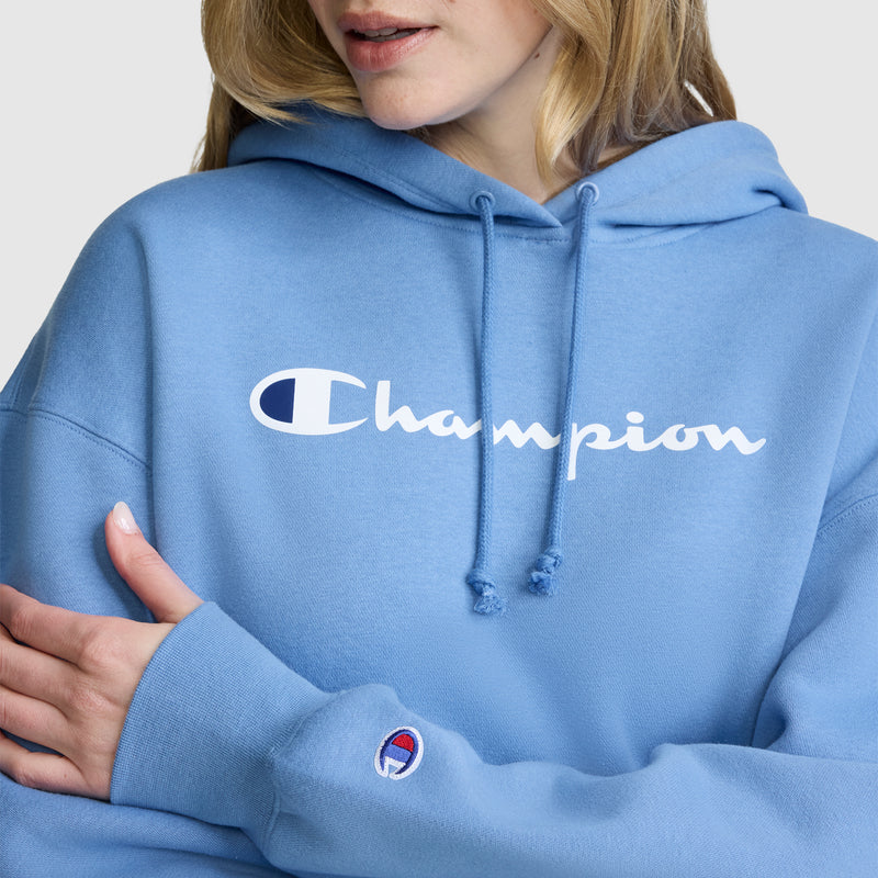 A person wearing a light blue Champion Women’s Powerblend Relaxed Hoodie, featuring the white script logo across the chest and a logo patch on the sleeve, stands with arms crossed in a relaxed pose, only the lower half of their face visible.