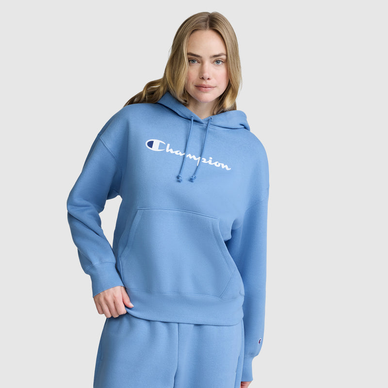 A woman wearing a light blue Champion Women’s Powerblend Relaxed Hoodie with a script logo and matching sweatpants, both crafted from cozy Powerblend fleece, stands against a plain white background.