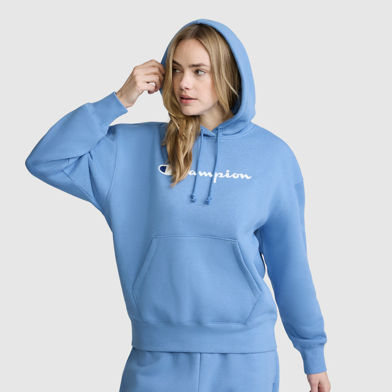 A woman wearing a light blue Champion Women’s Powerblend Relaxed Hoodie with a script logo stands against a plain background, touching her hair with one hand and looking to the side.