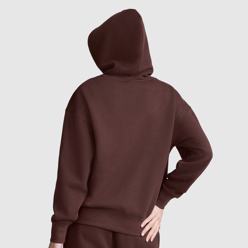 A person, seen from behind with one hand on their hip, models a brown Champion Women’s Powerblend Relaxed Hoodie Script Logo against a plain light gray background.