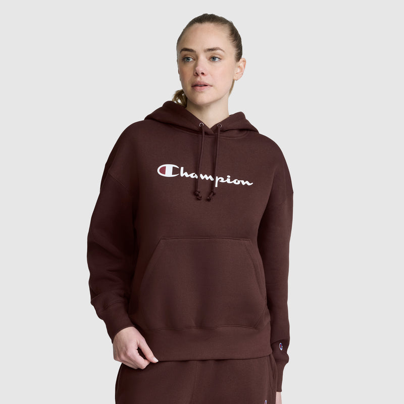 A woman wearing a brown Champion Women’s Powerblend Relaxed Hoodie, Script Logo, and matching sweatpants stands against a plain gray background, looking slightly to the side with one hand in her pocket. The soft Powerblend fleece of this Champion hoodie brings both comfort and style to her relaxed look.