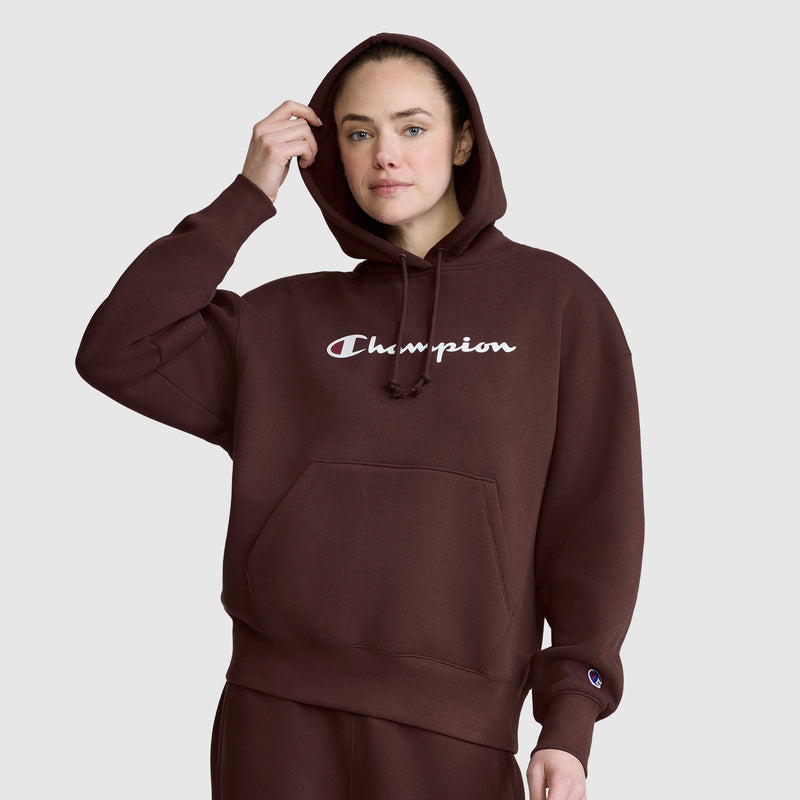 A person wearing a brown Champion Women’s Powerblend Relaxed Hoodie, Script Logo stands against a plain light gray background, touching the hood with one hand.