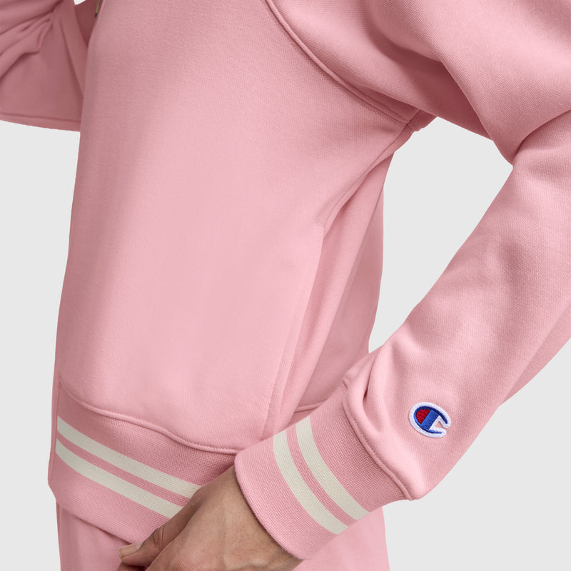 A person wears a pink Champion Pitcher Full-Zip Mockneck Top, C Logo, made of soft French terry with white stripes on the cuffs and waistband. A small blue, red, and white Champion logo patch accents the sleeve. The background is plain and light-colored.