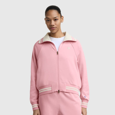 A woman stands facing forward against a light gray background, wearing a Champion Pitcher Full-Zip Mockneck Top with C Logo in light pink with a cream collar and stripes, made from soft French terry, paired with matching pink pants.