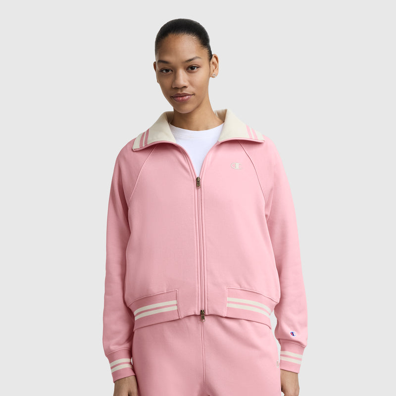 A woman stands facing forward against a light gray background, wearing a Champion Pitcher Full-Zip Mockneck Top with C Logo in light pink with a cream collar and stripes, made from soft French terry, paired with matching pink pants.