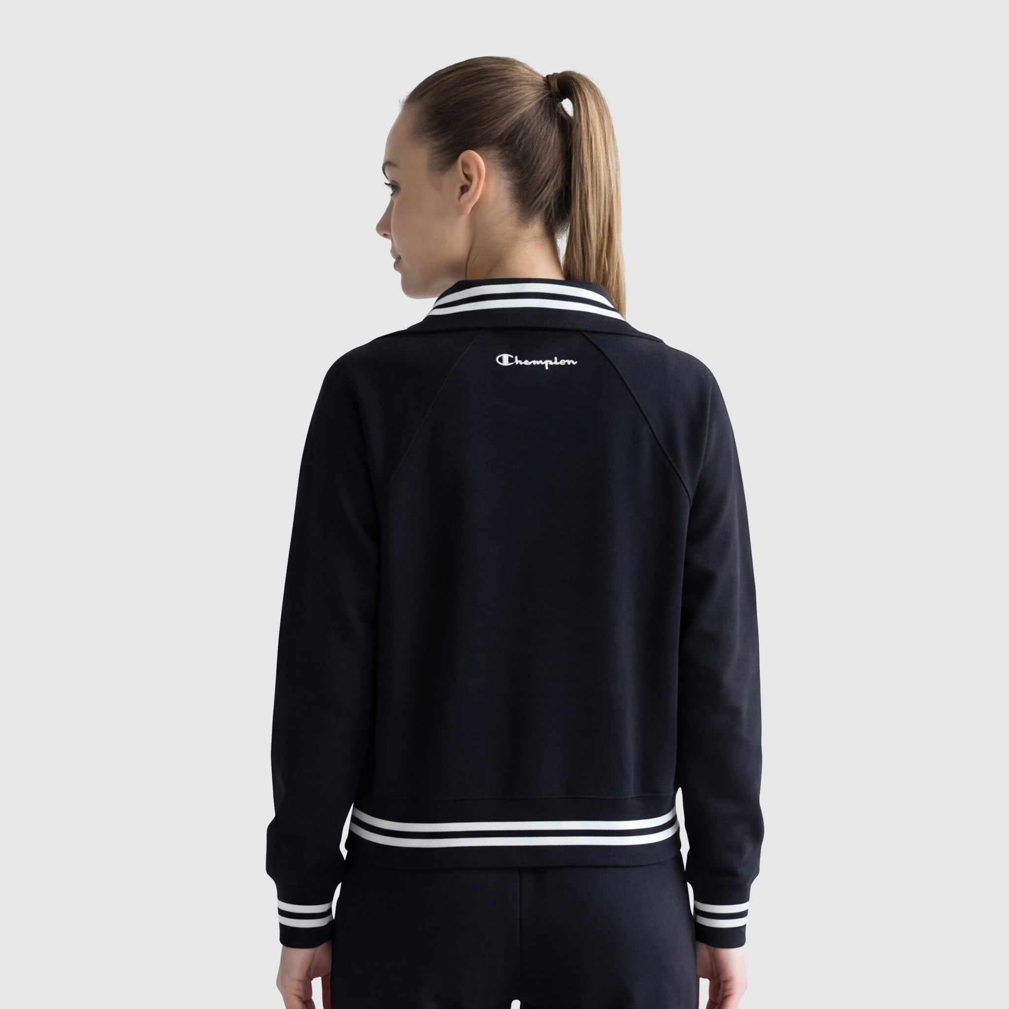 A woman with light skin and brown hair in a ponytail is seen from the back, wearing a black Champion Pitcher Full-Zip Mockneck Top with C Logo and white trim, paired with matching pants. The background is light gray.