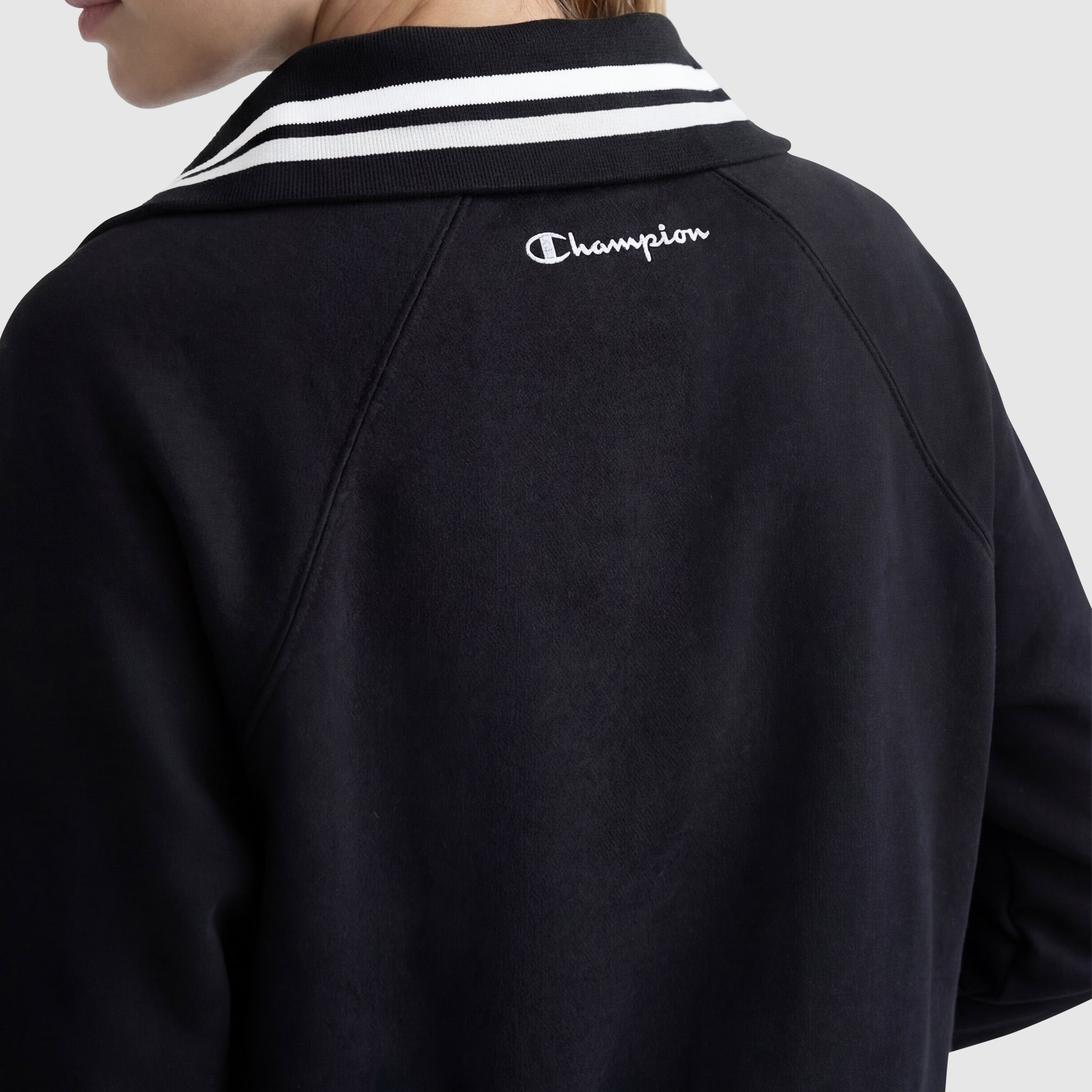 A person wears the Champion Pitcher Full-Zip Mockneck Top, C Logo, in black French terry with a white striped collar and white C logo on the upper back. The photo is taken from behind, showing part of their face and shoulder.