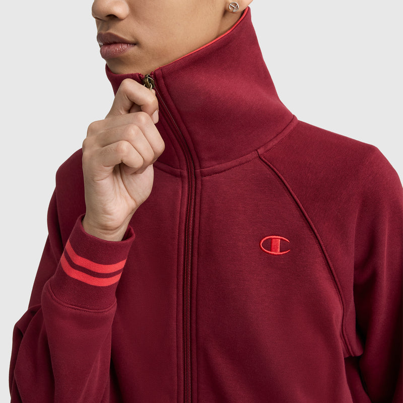 A person zips up a maroon Champion Pitcher Full-Zip Mockneck Top with red-striped cuffs and the C Logo on the chest, set against a light gray background.