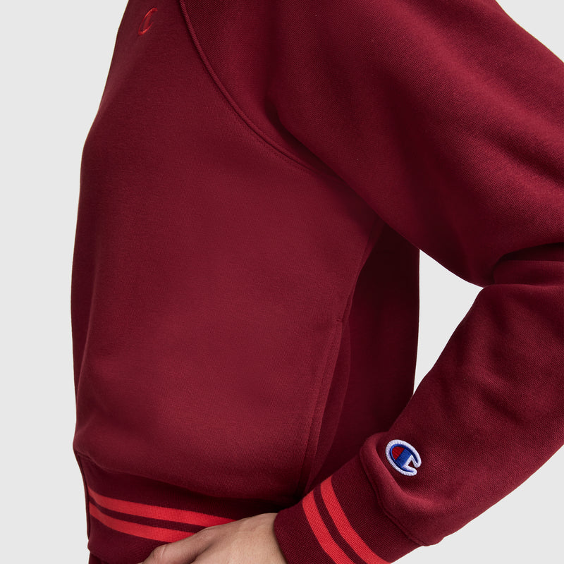 A person wears a maroon Champion Pitcher Full-Zip Mockneck Top with C Logo, made from soft French terry and detailed with red and maroon striped cuffs and waistband; only the torso and part of one arm show against a gray background.