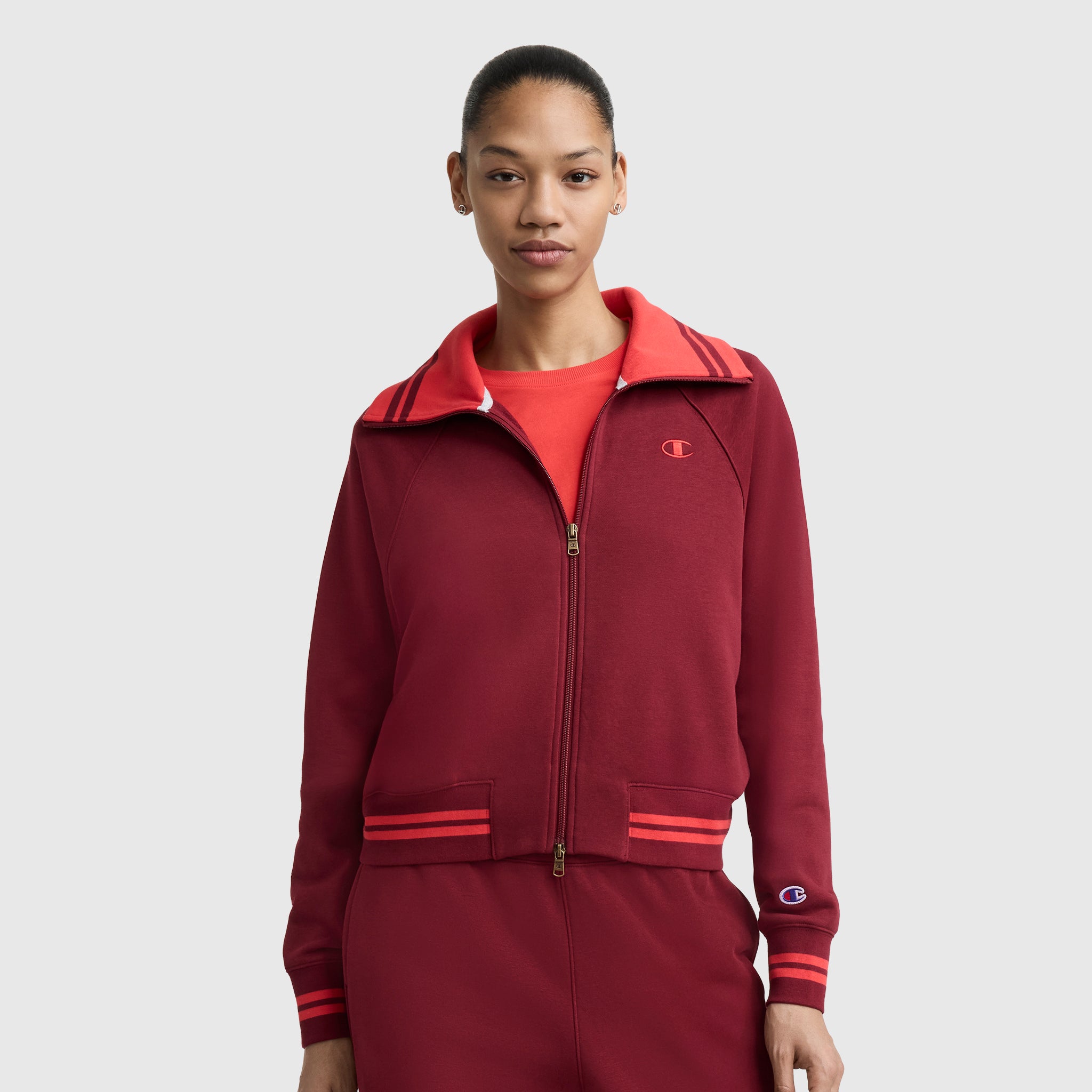 A person wears a matching maroon Champion Pitcher Full-Zip Mockneck Top with C Logo and pants, featuring red shoulder stripes. They stand against a plain light gray background.