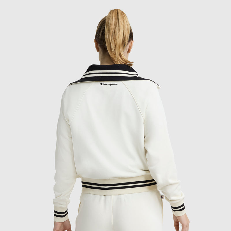 A person with blonde hair in a ponytail is seen from the back, wearing an off-white Champion Pitcher Full-Zip Mockneck Top featuring a C logo on the collar and matching pants with black trim.
