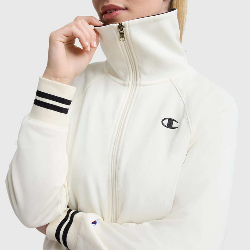 Wearing the Champion Pitcher Full-Zip Mockneck Top with black cuff stripes and a C logo on the chest, a person stands against a plain background, raising their hand to partially cover their mouth.