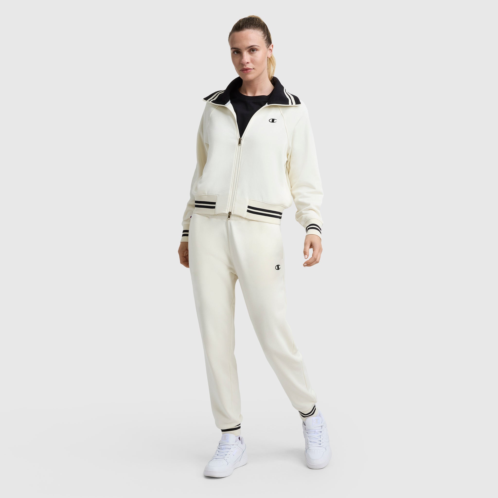 A person wears the Champion Pitcher Full-Zip Mockneck Top with C Logo in cream, paired with matching track pants featuring black and white stripes, a black shirt underneath, and white sneakers, standing against a plain light background.