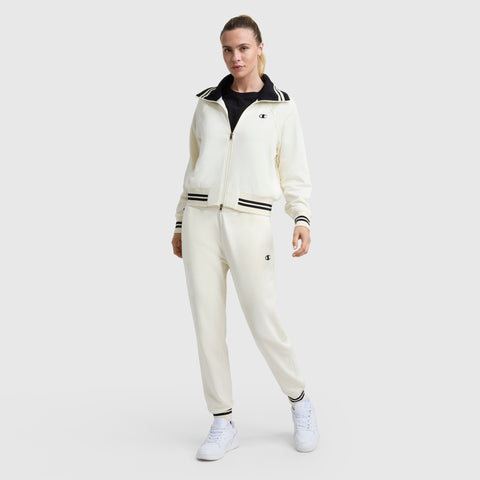 A person wears the Champion Pitcher Full-Zip Mockneck Top with C Logo in cream, paired with matching track pants featuring black and white stripes, a black shirt underneath, and white sneakers, standing against a plain light background.