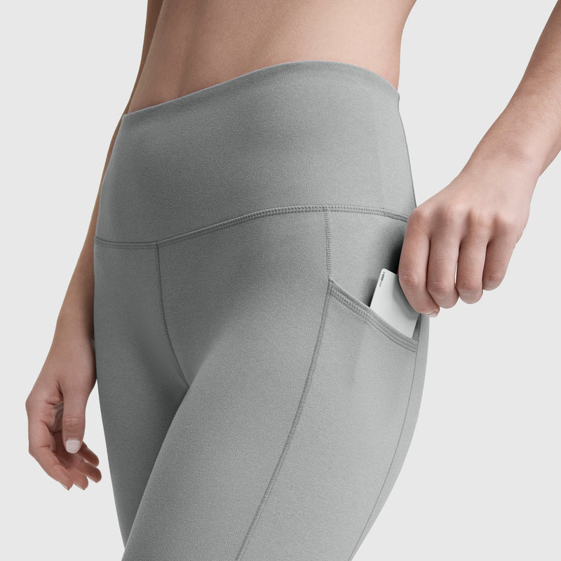 A person wearing Champion Leggings, C Logo, slips a white smartphone into the side pocket. The plain background highlights the leggings’ silky softness, stretch fit, and convenient pocket feature.