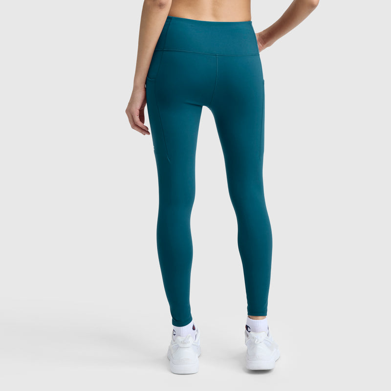 A person is shown from the back, standing against a plain light background, wearing silky soft teal Champion Leggings, C Logo, paired with white sneakers.