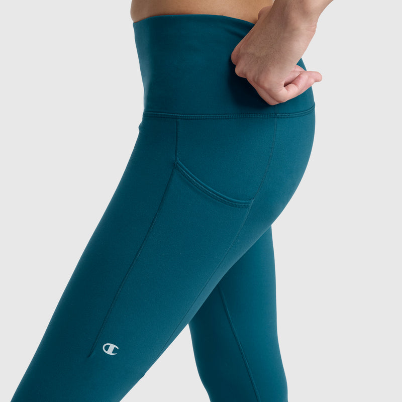 A person wearing Champion C Logo Leggings in teal, featuring a small Champion logo on the left thigh and a high-rise waistband. Their right hand rests near the waistband, highlighting the side pocket and functional, stylish design. The background is plain light gray.