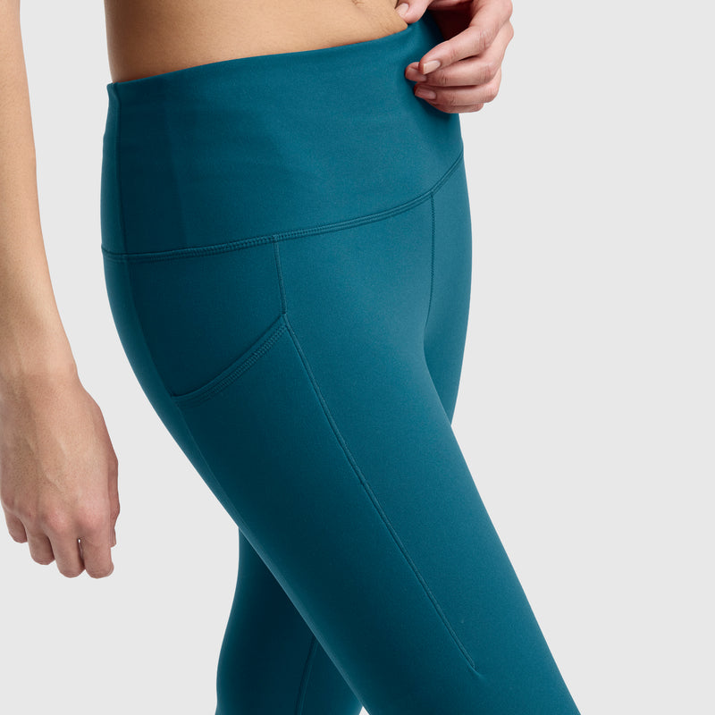 A person wearing Champion’s Leggings, C Logo in teal is shown from the waist down, with one hand resting on their hip. These high-waisted, silky soft leggings feature a side pocket and visible seam details, making them perfect for the studio.