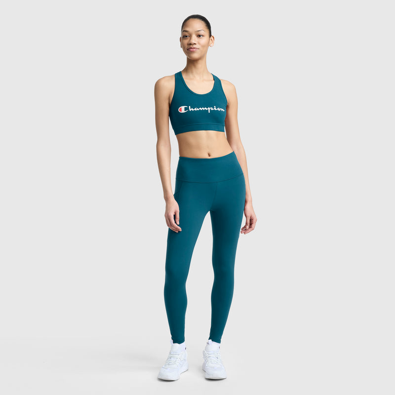 A person stands facing forward wearing teal Champion C Logo leggings with pockets and a matching sports bra, paired with white sneakers against a plain light background.