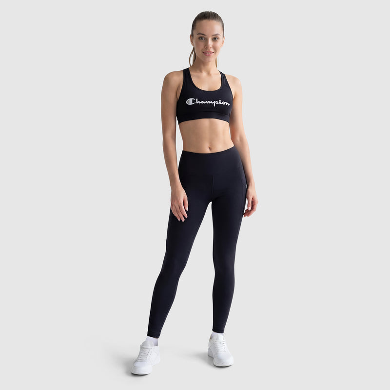 A woman models black Champion Leggings, C Logo with side pockets and a matching black Champion sports bra, smiling confidently against a light grey background.