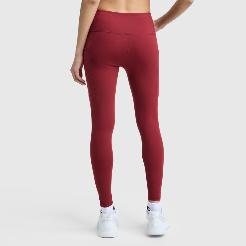 A person is shown from the back wearing Champion C Logo high-waisted, red leggings with side pockets and white sneakers, standing against a plain white background with one hand on their hip.