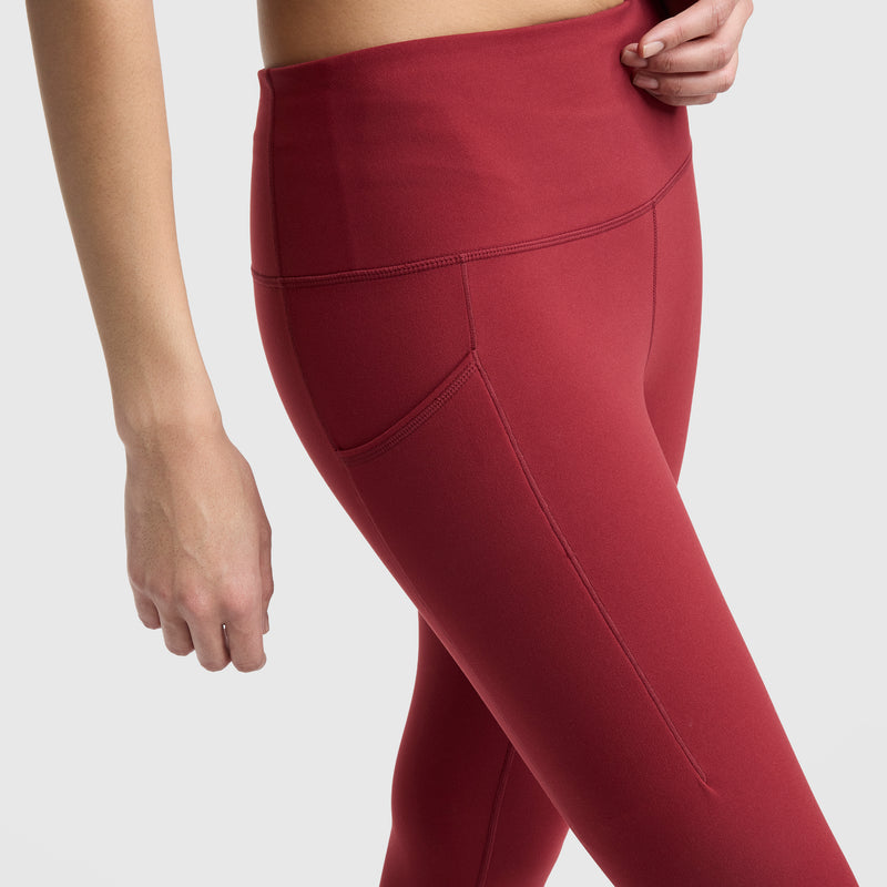 A person wears Champion’s high-waisted C Logo leggings in red, made from peached fabric with side pockets; only their lower torso and upper legs are shown against a plain light background.
