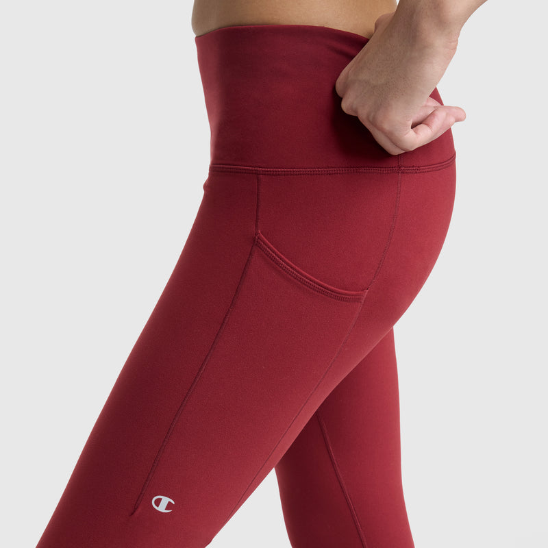 Person wearing high-waisted, red Champion C Logo leggings with side pockets and visible seams. The soft peached fabric stands out against a plain light gray background, adding a simple yet stylish touch. One hand rests on their hip.