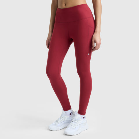 A person wears high-waisted red Champion C Logo leggings with side pockets and white athletic shoes, standing against a plain light gray background. The upper body is partially out of frame.
