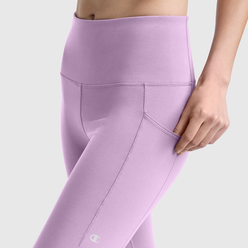 A person wearing Champion Leggings, C Logo—high-waisted, lavender leggings with pockets crafted from silky soft, peached fabric—rests one hand on the side pocket and is shown from mid-torso to mid-thigh against a plain background.
