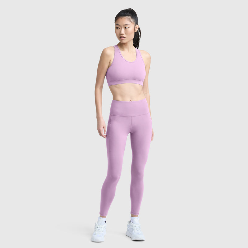 A woman stands against a plain light gray background, facing slightly to the side with her hands by her sides, wearing Champion’s C Logo leggings in light purple with pockets and a matching sports bra. She completes the look with white sneakers and a ponytail hairstyle.