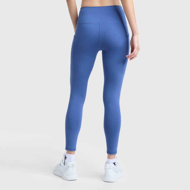 A person faces away, wearing Champion C Logo high-waisted blue leggings with side pockets and white sneakers, set against a plain light gray background.