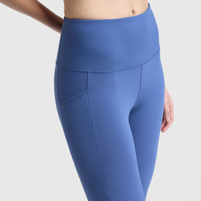 A person wearing Champion C Logo high-waisted blue leggings with side pockets, crafted from soft peached fabric, is shown from mid-torso to mid-thigh against a plain light background.