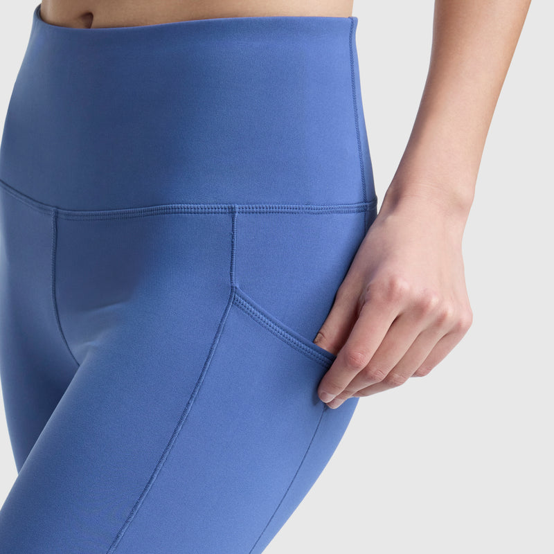 A person wears Champion's C Logo high-waisted blue leggings, crafted from soft peached fabric, and places their hand in the convenient side pocket. The plain, light background accentuates the leggings.
