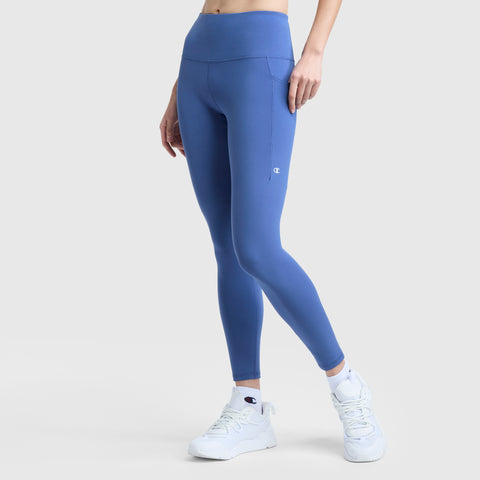 Someone stands against a light grey background wearing Champion Leggings, C Logo—high-waisted blue leggings with side pockets and a small logo on the upper thigh—paired with white athletic sneakers.