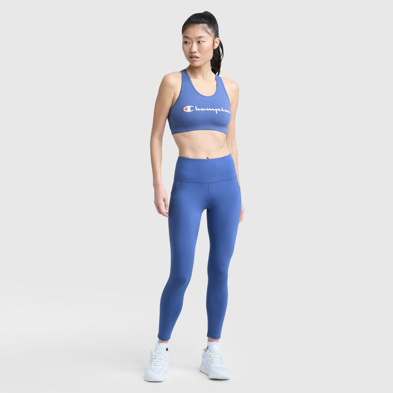 A woman in Champion’s C Logo leggings and a matching blue sports bra stands against a white background, looking to the side. Her long hair is tied back and she wears white sneakers. The high-waisted leggings feature side pockets.