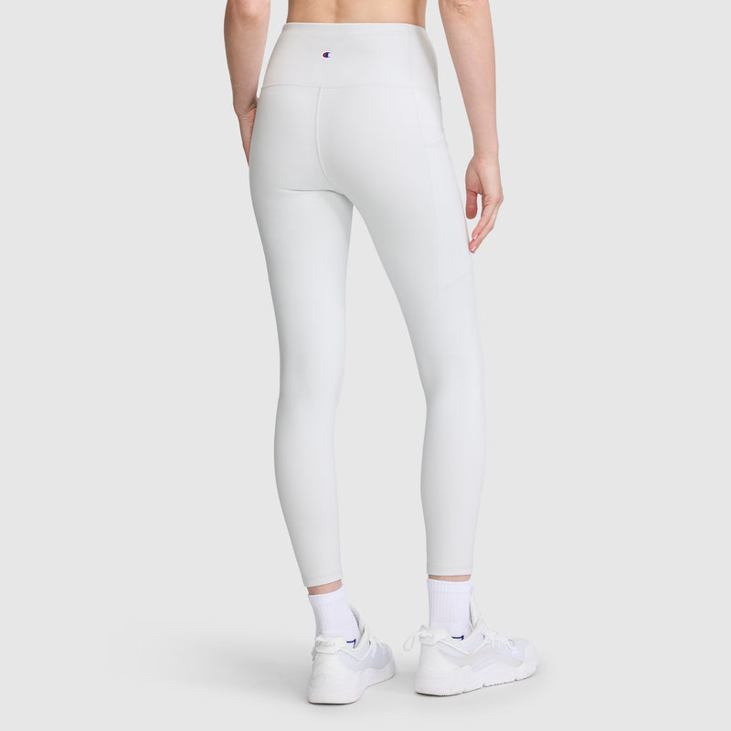A person is wearing Champion’s Leggings, C Logo—high-waisted leggings crafted from soft peached fabric with side seams, visible stitching, discreet side pockets, and a small Champion logo on the waistband—paired with white sneakers and socks, standing against a plain light background.